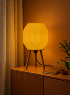Lampe à poser Tripod beige Luciole allumée, avec abat-jour strié imprimé en 3D et trois pieds en bois, diffusant une lumière douce et chaleureuse.